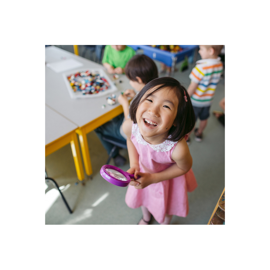 A young child wearing a pink dress, holding a magnifying glass, looking at the camera and smiling.